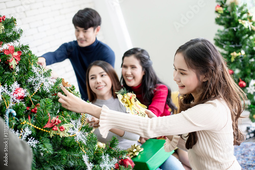 A beautiful Asian woman is setting up a Christmas tree, Smiling. Her friends are decorating the Christmas tree in the background.