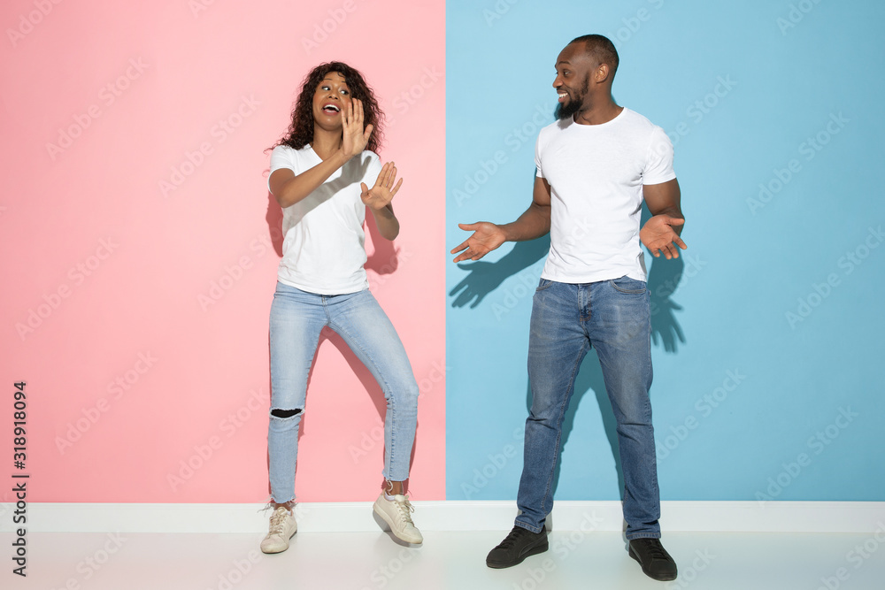 Fototapeta premium She's having fun. Young emotional man and woman in casual clothes on pink and blue bicolored background. Concept of human emotions, facial expession, relations, ad. Beautiful african-american couple.