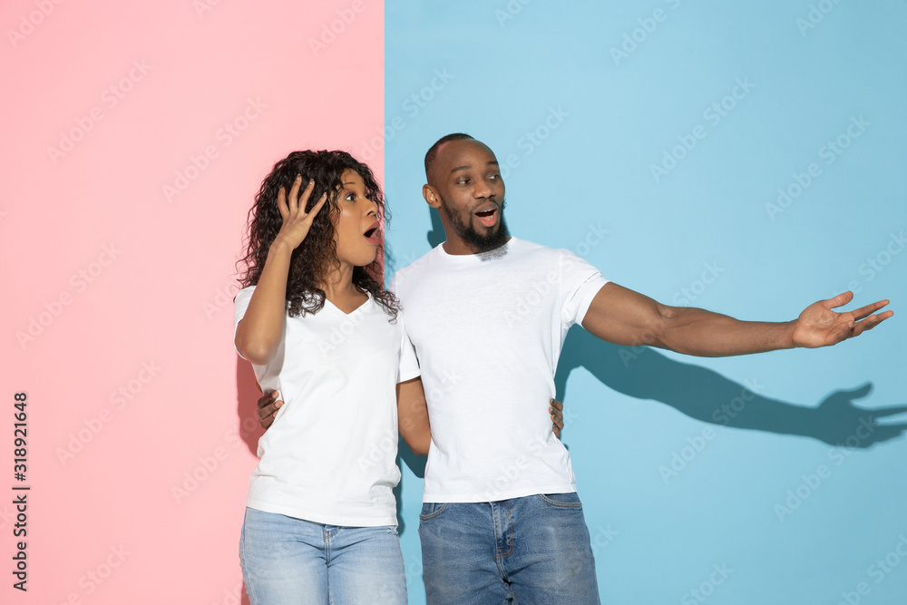 Fototapeta premium Pointing together. Young emotional man and woman in casual clothes on pink and blue bicolored background. Concept of human emotions, facial expession, relations, ad. Beautiful african-american couple.