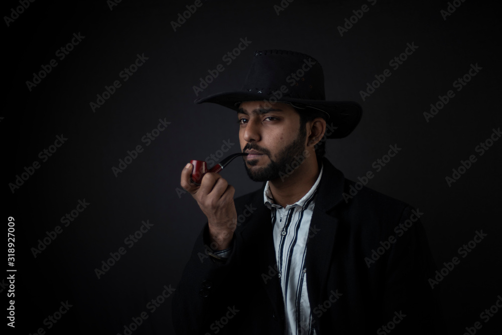 Fashion portrait of an young and handsome Indian Bengali brunette man with striped formal shirt, black suit, hat and a pipe in black copy space background. Indian lifestyle and fashion photography.