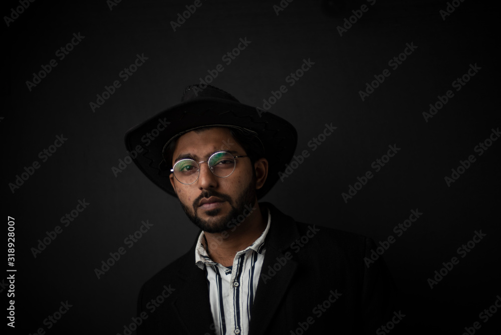Fashion portrait of young and handsome Indian Bengali brunette man with striped formal shirt, black suit, glasses and cowboy hat in black copy space background. Indian lifestyle and fashion photograph