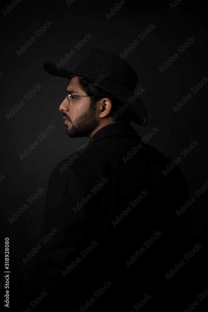 Fashion portrait of young and handsome Indian Bengali brunette man with striped formal shirt, black suit, glasses and cowboy hat in black copy space background. Indian lifestyle and fashion photograph