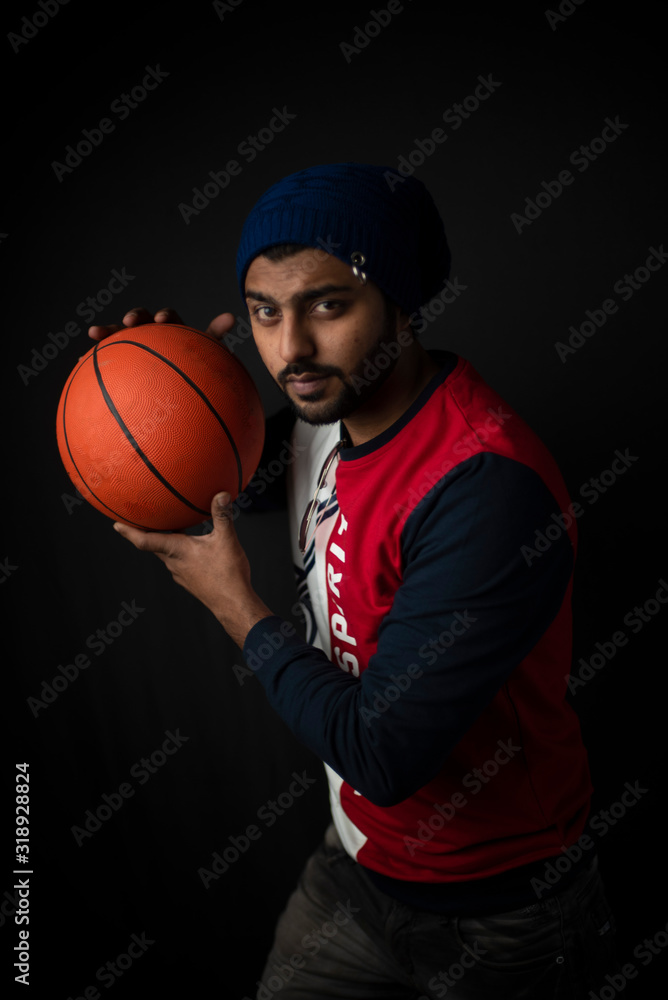 Fashion portrait of an young Indian Bengali brunette man in casual tee shirt and head band playing with basket ball in black copy space background. Indian lifestyle and fashion photography. Fashion portrait of an young Indian Bengali brunette man in casual tee shirt and head band playing with basket ball in black copy space background. Indian lifestyle and fashion photography.