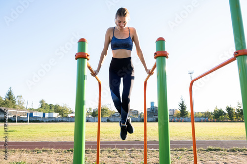Young sportive athlete woman doing fitness exercise on metal bars outdoors at stadium court.