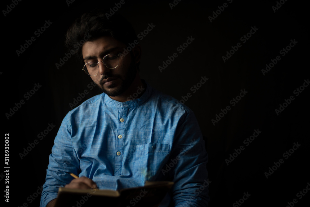 Fashion portrait of young Indian Bengali brunette man with traditional cotton wear and glasses holding pencil and book in black copy space background. Indian lifestyle and fashion photography Fashion portrait of young Indian Bengali brunette man with traditional cotton wear and glasses holding pencil and book in black copy space background. Indian lifestyle and fashion photography