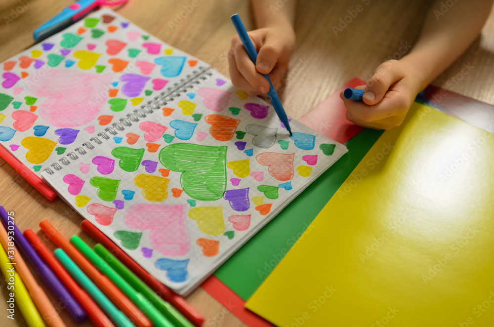 Child's hands draw hearts of different colors in a notebook. Close-up photo. Child's hands draw hearts of different colors in a notebook. Close-up photo.