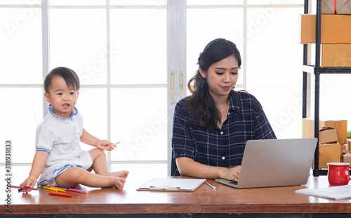 Beautiful Asian business mom is using laptop, smiling, spending time with cute baby girl at home, Single mother take care adorable child kid, working on technology computer cardboard box background