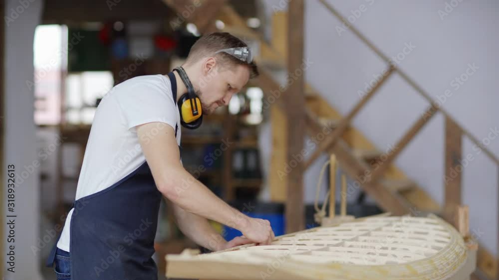 Side view medium shot of focused carpenter building large wooden model of boat standing at workbench in carpentry shop Side view medium shot of focused carpenter building large wooden model of boat standing at workbench in carpentry shop