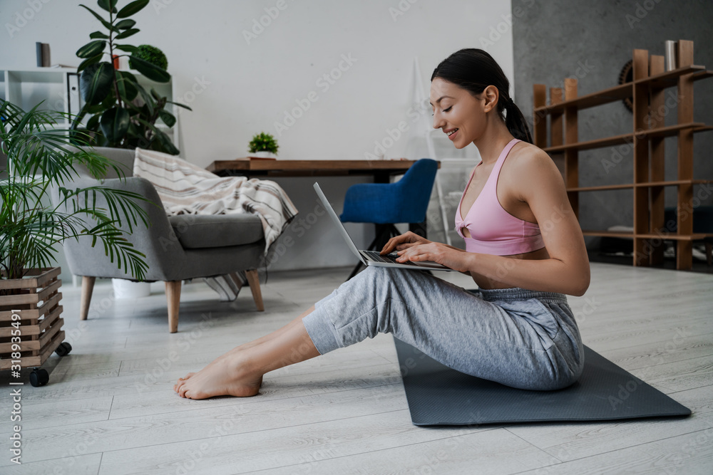Young athletic millennial girl sits on the floor in her apartment on sports mat before morning exercises, searches online for exercises on certain muscle groups, stretching and yoga asanas on laptop Young athletic millennial girl sits on the floor in her apartment on sports mat before morning exercises, searches online for exercises on certain muscle groups, stretching and yoga asanas on laptop