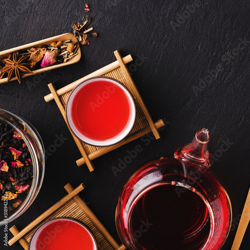 Aromatic fresh brew natural red fruit tea in two bowls ,glass teapot,glass jar and plate with dry tea leaves on a black concrete background