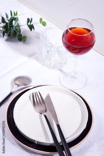 Tableware and decorations for serving a festive table. Plates, red wine glass and cutlery with green leaves on white textile background.