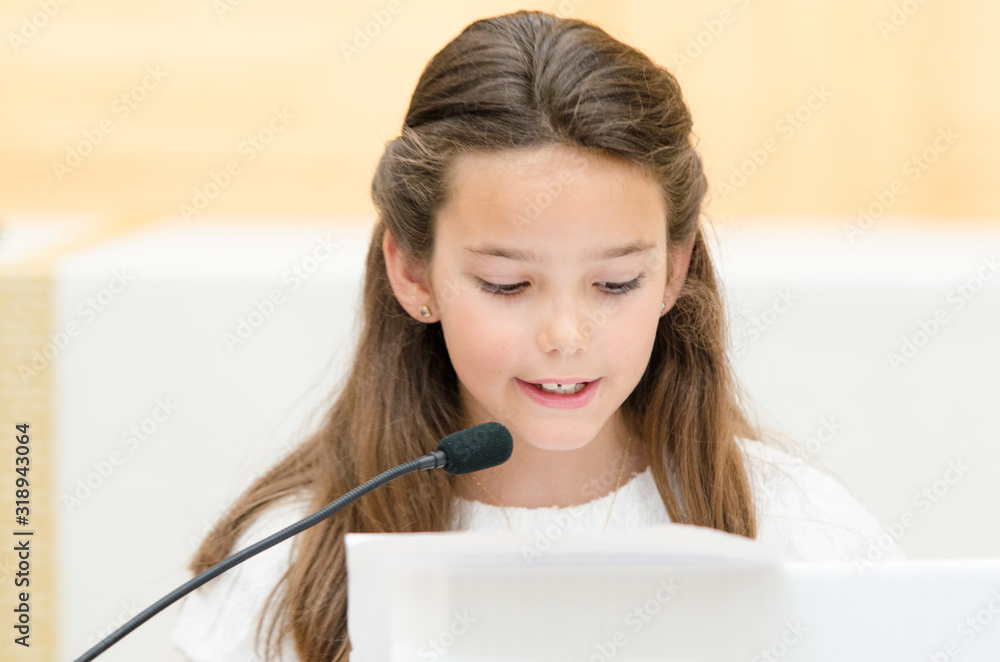 Little girl saying a few words in front of the microphone on the day of her first communion. Little girl praying at the microphone