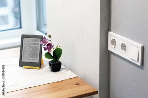 Group of white european electrical outlets and a switch located on a gray wall in a light modern kitchen with flower and e-book on the table by the window. Selective focus