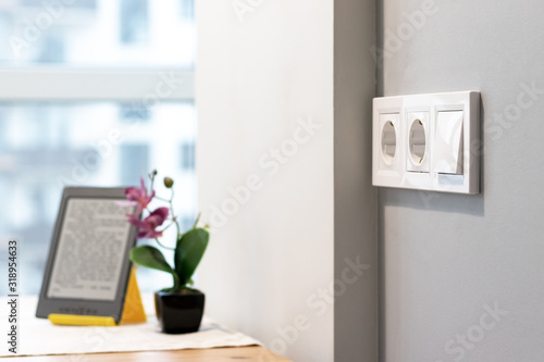 Group of white european electrical outlets and a switch located on a gray wall in a light modern kitchen with flower and e-book on the table by the window. Selective focus