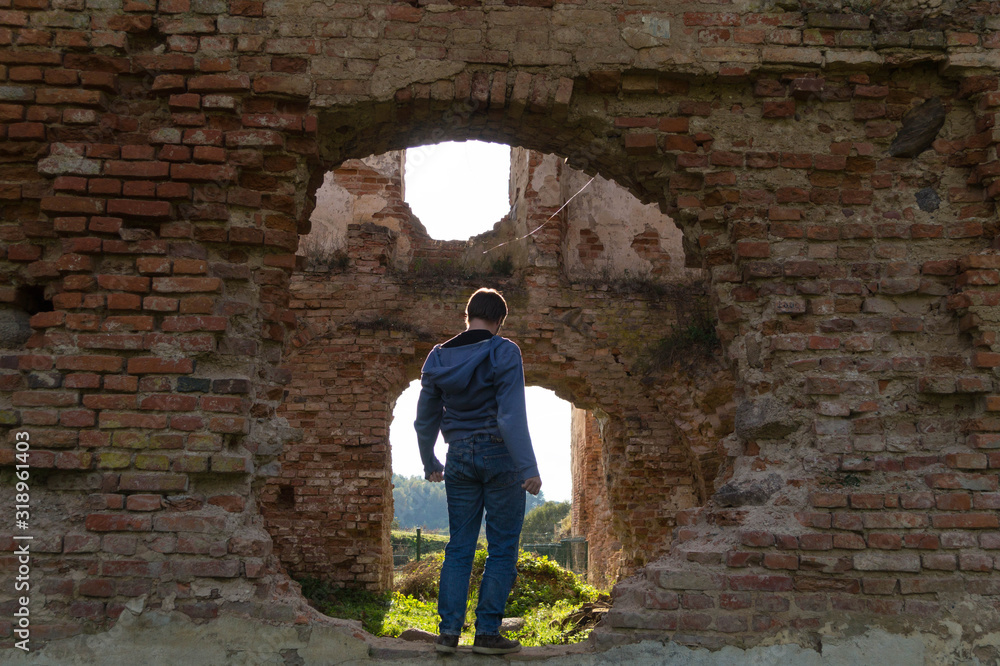 A young man in blue clothes stands on the ruins of an old Golshany castle, turning his back, framed by a round hole in the castle wall
