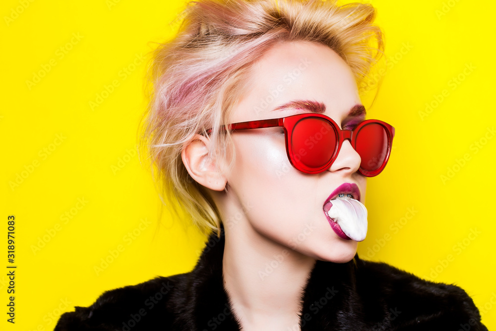 Portrait of a blonde in red glasses on a yellow background with chewing gum in her mouth. A woman is chewing gum. Portrait of a blonde in red glasses on a yellow background with chewing gum in her mouth. A woman is chewing gum.