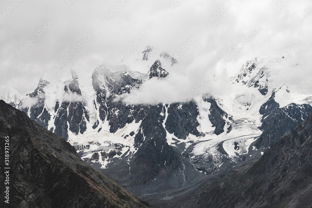 Obraz premium Atmospheric alpine landscape with massive hanging glacier on giant mountain. Big glacier tongue on mountainside. Low clouds among snowbound mountains. Cracks on ice. Majestic scenery on high altitude.