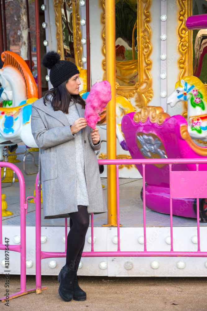 General shot of young brunette woman, with black cap, looking to the right, with pink cotton candy, in front of a carousel, with sunset light, vertically, in Madrid, Spain