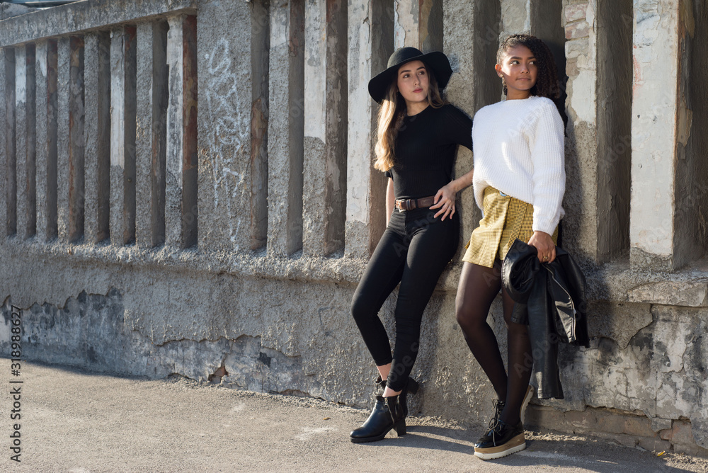 general plan of two beautiful young Spanish girls, generation Y, are standing at the old texture wall, smiling general plan of two beautiful young Spanish girls, generation Y, are standing at the old texture wall, smiling