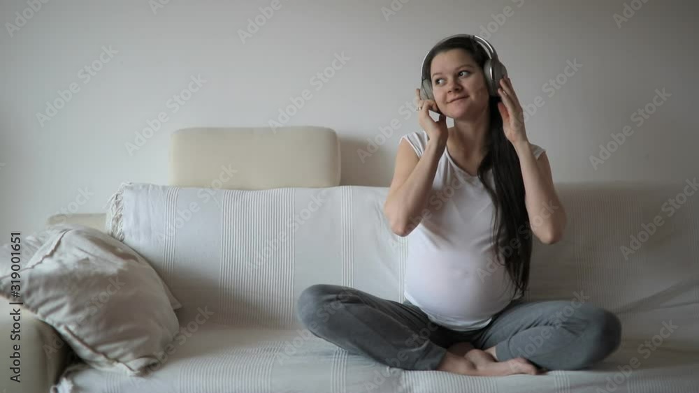 Pregnant woman dancing on white sofa at home and listening music in headphones Pregnant woman dancing on white sofa at home and listening music in headphones