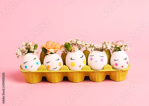 Easter creative concept. Easter decorative eggs with painted cheerful faces and flowers instead of hair in the cardboard egg tray on the pink backgraund. Copy space