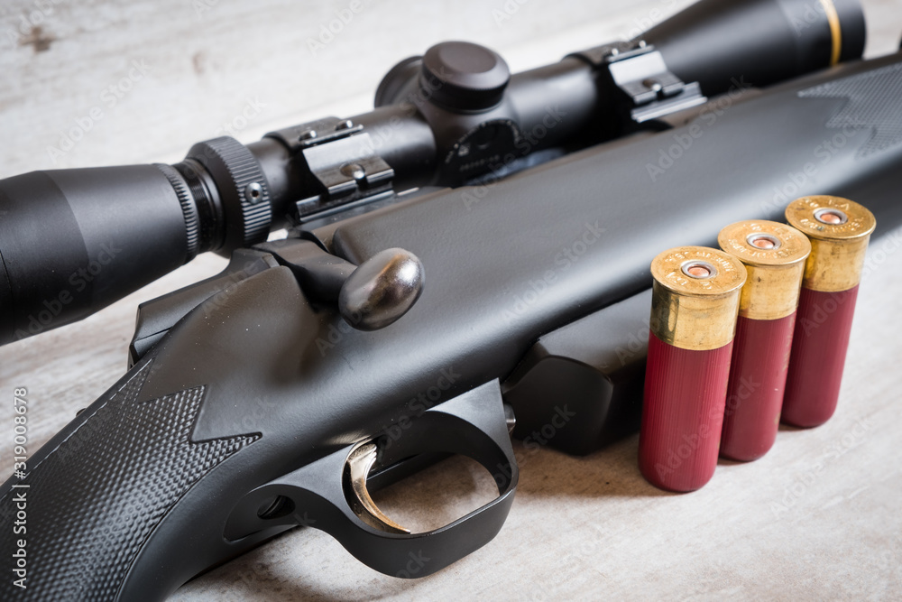 Obraz premium A bolt action scoped shotgun and three shotgun shells against a light wooden background. The firearm is often mistaken for a rifle due to its hybrid construction, bolt, and use of high powered scope