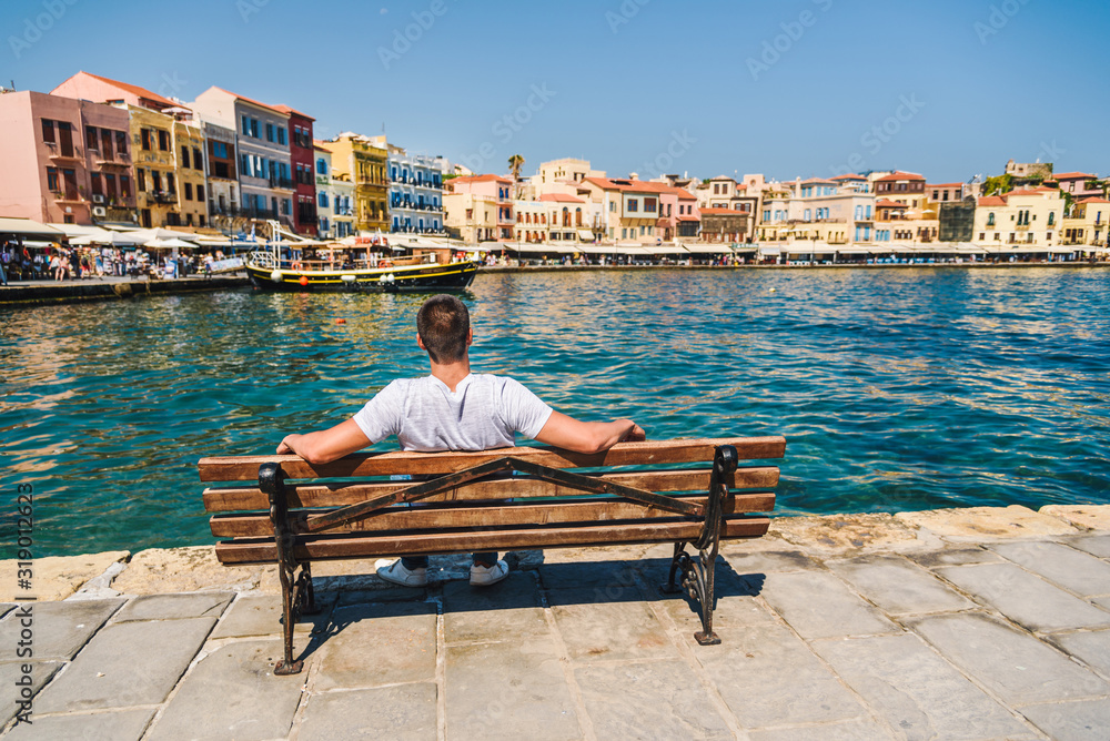 Young male tourist sitting on a bench and looking at the port of Chania, Crete, Greece. Tourist concept. Sightseeing. Summer vacation destination. Travel concept
