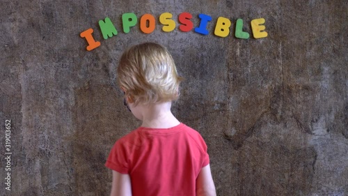 funny blond boy in red clothes is standing against wall where word is written: impossible. Boy does not like this word and he covers first two letters with his palms in order to succeed: possible