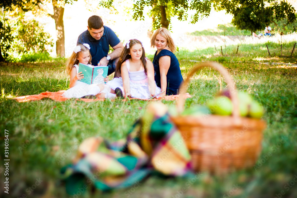 Happy Two Parent Family With Two Daughters On Orange Picnic Blanket In The Park. Picnic basket in bokeh