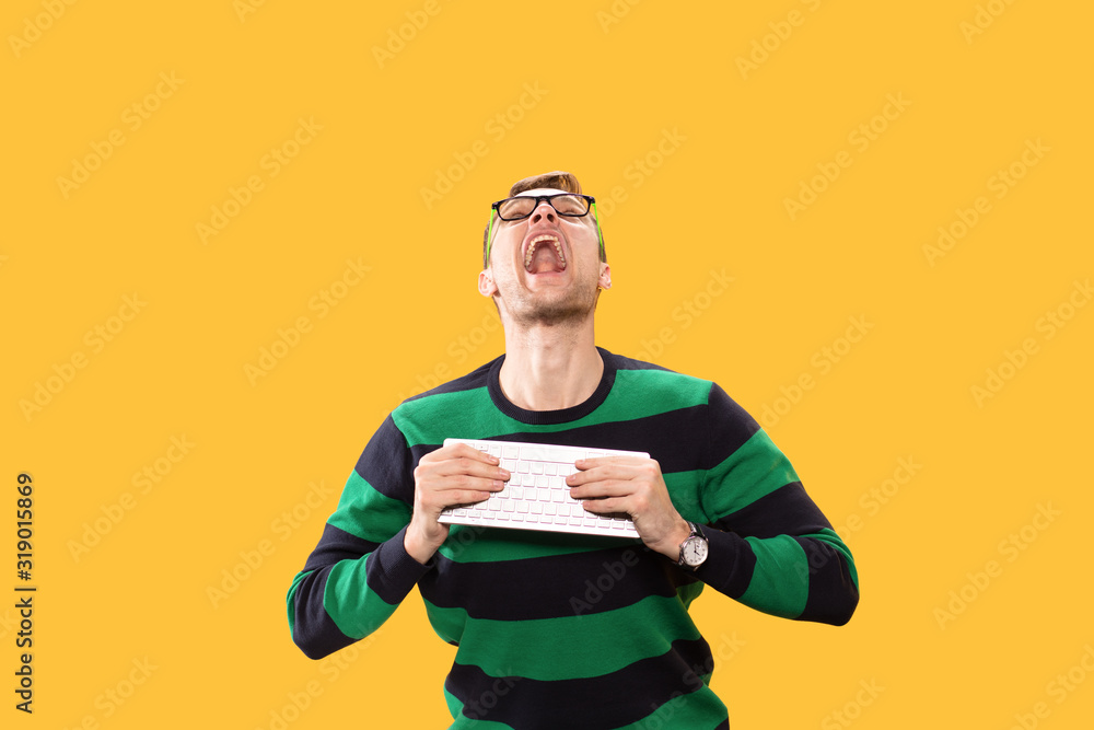 Angry young man holding a keyboard in his hands and trying to break it on yellow background. He screams heavily and looks up Studio photo.