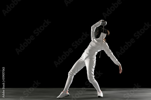 Catching moment. Teen girl in fencing costume with sword in hand isolated on black background. Young female model practicing and training in motion, action. Copyspace. Sport, youth, healthy lifestyle. © master1305