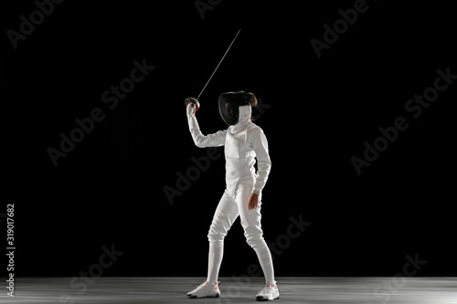 Graceful. Teen girl in fencing costume with sword in hand isolated on black background. Young female model practicing and training in motion, action. Copyspace. Sport, youth, healthy lifestyle.