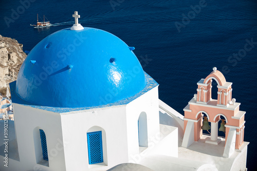 Blinding white Greek church featuring bright blue dome and pink bell tower against deep blue sea in Santorini, Greece