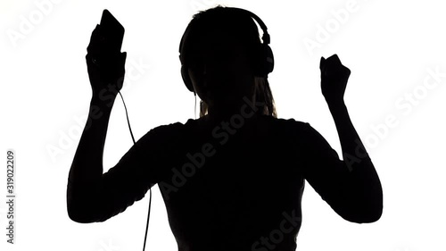 silhouette of a young happy woman listening to music in big headphones and holding a smartphone, girl dancing on white isolated background, concept lifestyle