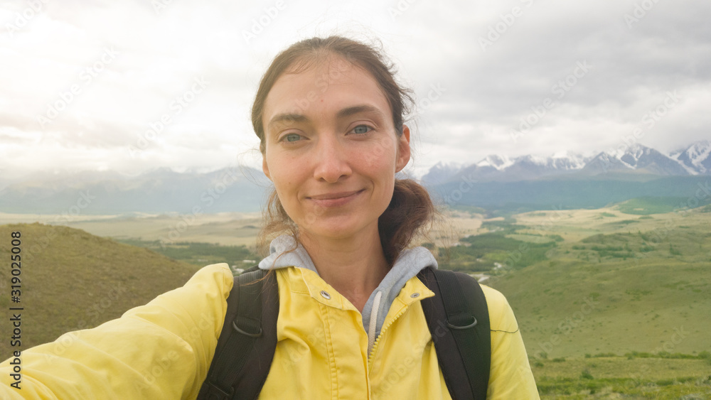 A brunette girl in a bright yellow jacket and with a backpack takes a selfie against the background of the beautiful Altai mountains.