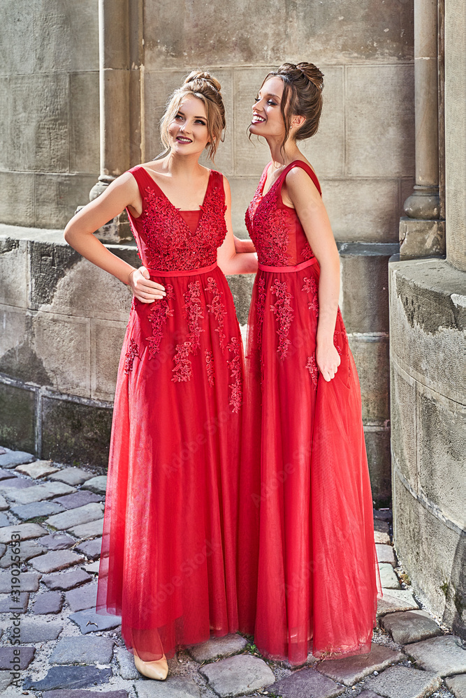 Two beautiful bridesmaids girls blonde and brunette ladies wearing elegant full length red chiffon bridesmaid dress with lace and holding flower bouquets. European old town location for wedding day.