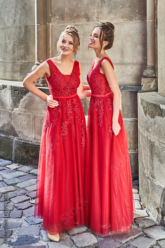Obraz na płótnie Two beautiful bridesmaids girls blonde and brunette ladies wearing elegant full length red chiffon bridesmaid dress with lace and holding flower bouquets. European old town location for wedding day. w salonie
