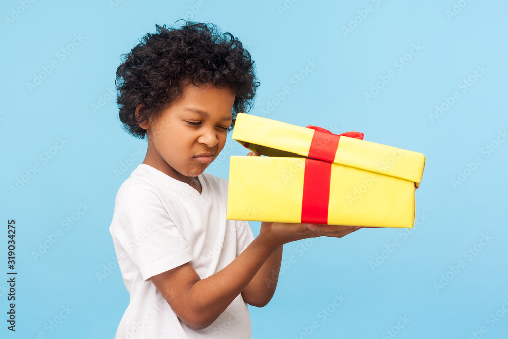 Portrait of curious adorable little boy peeking inside gift box, unpacking present with funny inquisitive look, impatient child unboxing birthday surprise. studio shot isolated on blue background - obrazy, fototapety, plakaty 