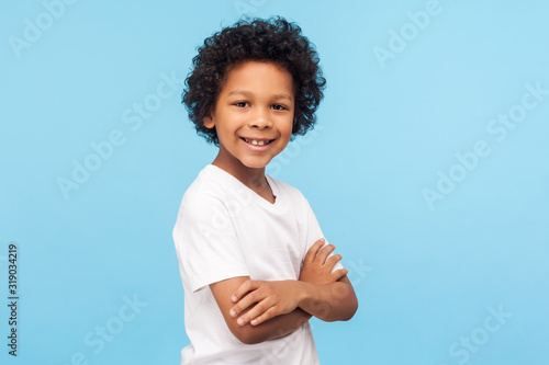 Portrait of happy and confident little boy in white T-shirt standing with crossed hands and smiling at camera, healthy cheerful child, positive emotions. indoor studio shot isolated on blue background