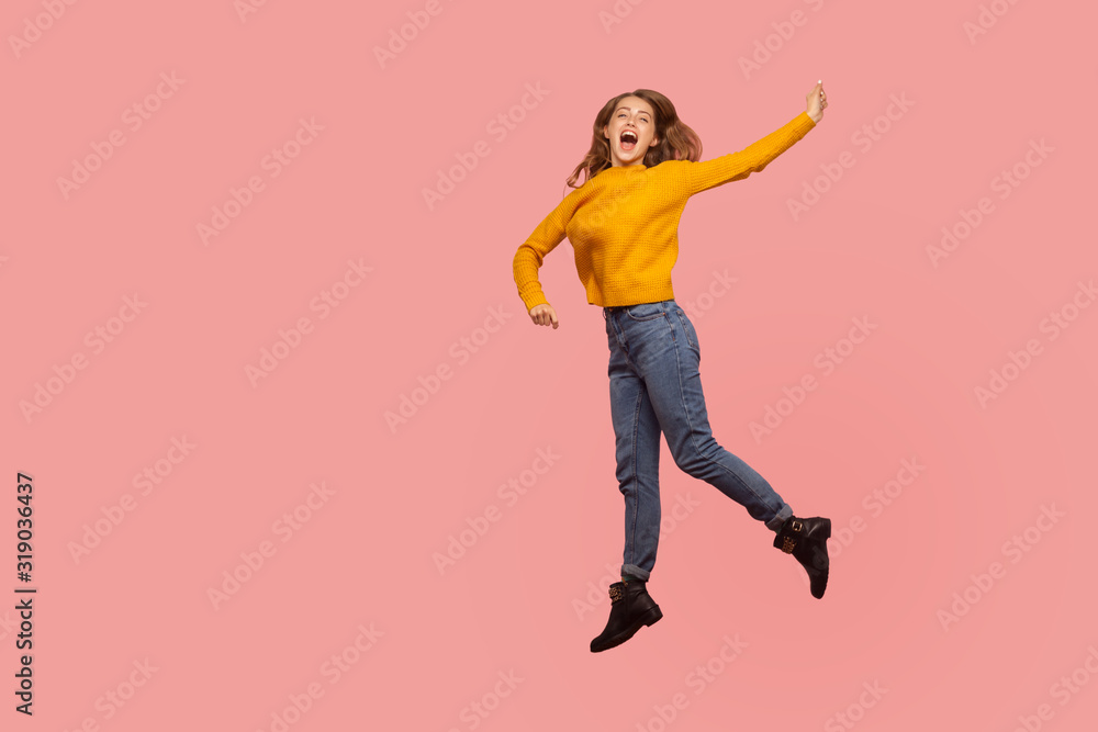 Portrait of lively energetic ginger girl in sweater and denim full of enthusiasm jumping in air or flying up, screaming from happiness. indoor studio shot isolated on pink background, copy space