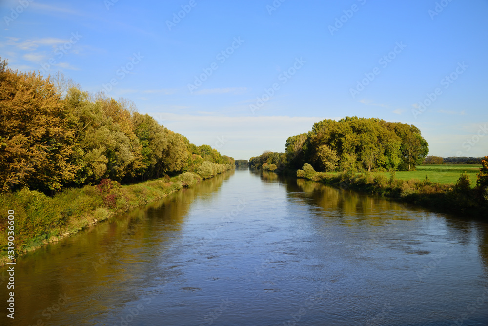 A large river flows calmly between an autumn landscape with trees and meadows with colorful leaves, under a blue sky. A large river flows calmly between an autumn landscape with trees and meadows with colorful leaves, under a blue sky.