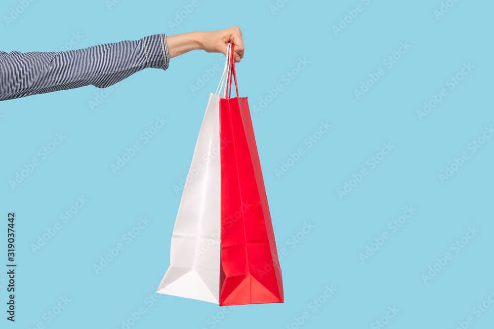 Closeup of shopping bags in woman arm, advertising of purchases in fashion store, female hand holding red and white packages, commercial concept. indoor studio shot isolated on blue background