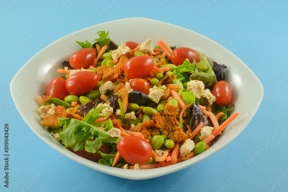 Healthy salad with fresh mixed greens, grape tomatoes, matchstick carrots, edamame, cheese and crispy red peppers in white bowl on blue background Healthy salad with fresh mixed greens, grape tomatoes, matchstick carrots, edamame, cheese and crispy red peppers in white bowl on blue background