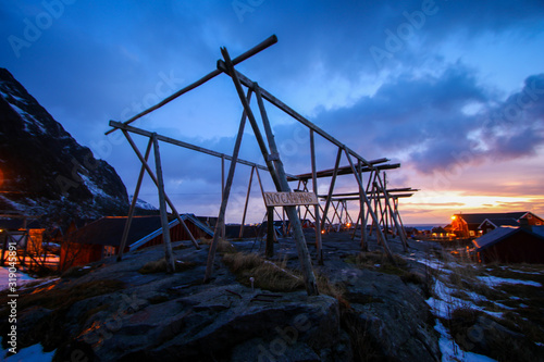 Wooden codfish drier in the small fishing village of A (Moskenes) at the end of the road of the Lofoten islands archipelago in northern Norway - Red rorbuer on stilts in winter at dawn in a fjord