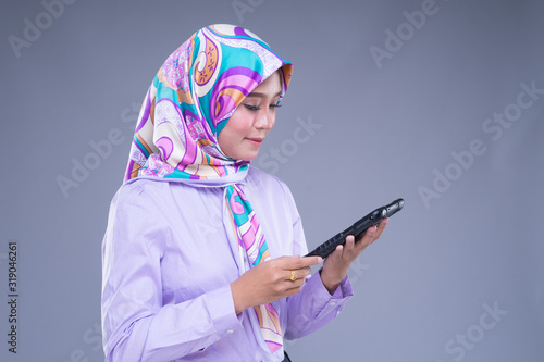 Half length portrait of an attractive Muslim woman wearing business attire and hijab poses holding a tablet isolated on grey background. For image cut-out for technology, business or finance. © HEMINXYLAN