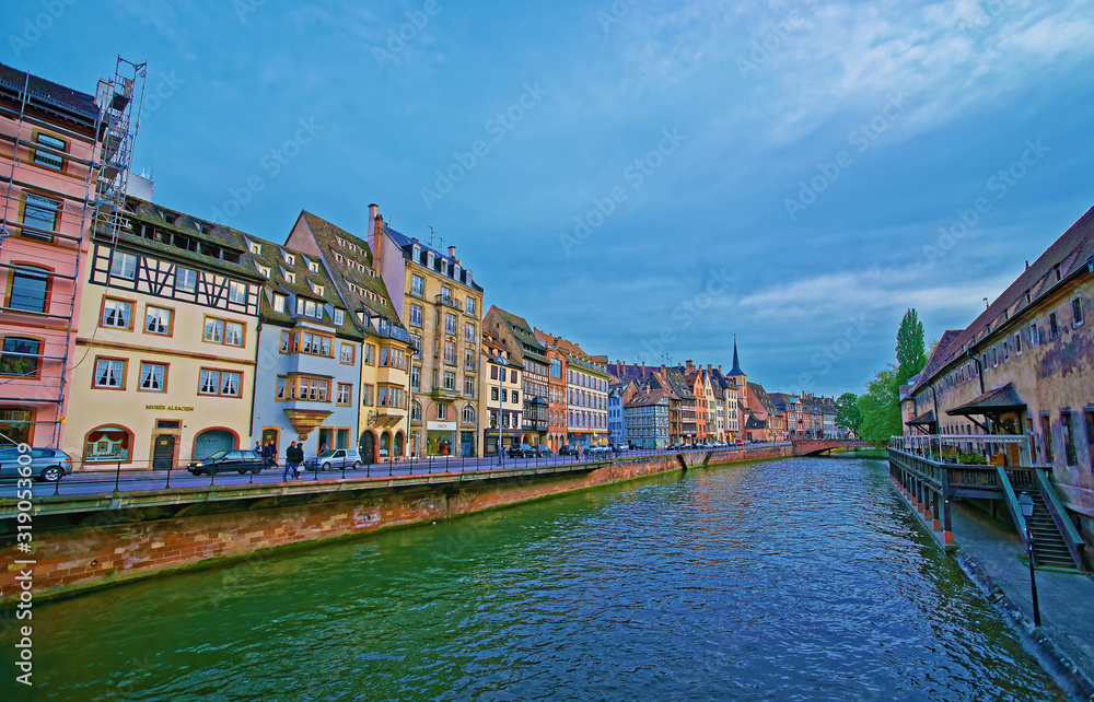 Strasbourg, France - April 30, 2012: Quay of Ill River with Spire of St Nicholas Church and Old Custom House in the old town of Strasbourg in Grand East region of France. People on the background