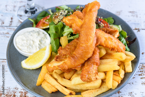 A plate of 'Fish and Chips' with battered fish, potato chips, salad, tartar sauce and lemon wedge.