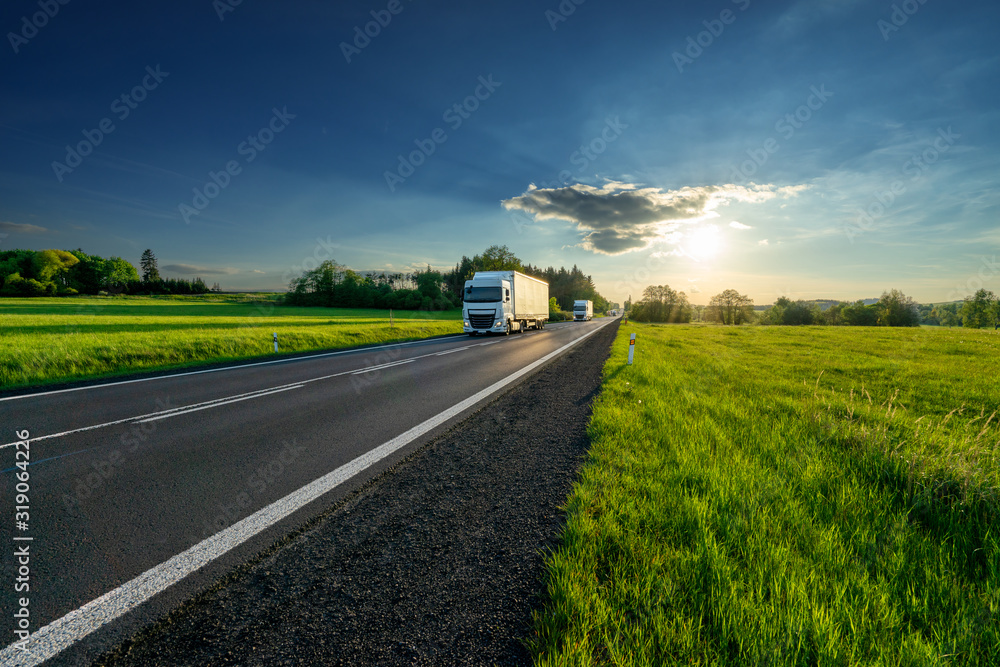 White trucks arriving from a distance on an asphalt road in rural landscape at sunset