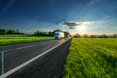 Obraz na płótnie White trucks arriving from a distance on an asphalt road in rural landscape at sunset w salonie