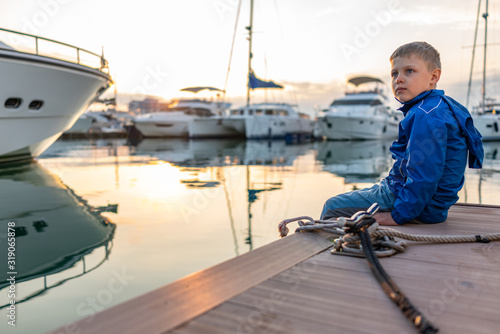 A boy in a blue jacket sits on a pier with yachts during sunset.